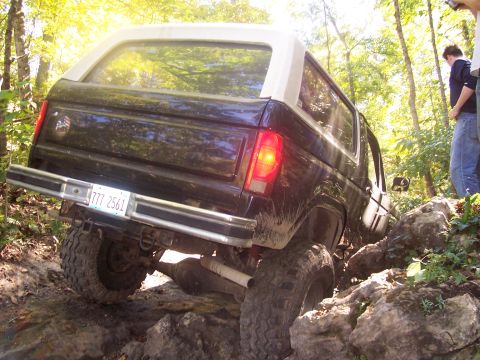 1986 Ford Bronco - Rear three-quarter view of a black two-door SUV with a white removable hardtop, crawling over large rocks on a wooded trail with a person s…