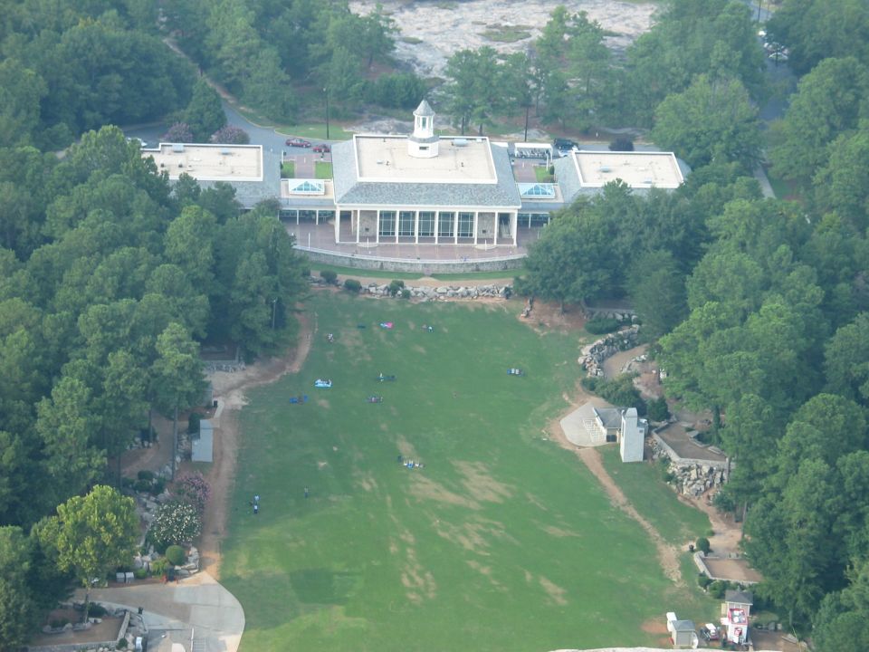 Aerial view of a large building with a colonnaded entrance, surrounded by trees and a large grassy lawn with people scattered across it