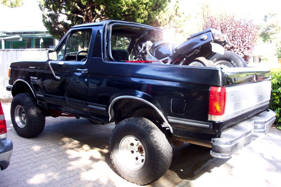 Black lifted Ford Bronco with a motorcycle loaded in the rear cargo bed, rear side view.
