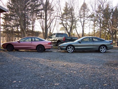 1996 Ford Probe - Two Ford Probes and a pickup truck are parked on a gravel lot with trees in the background.
