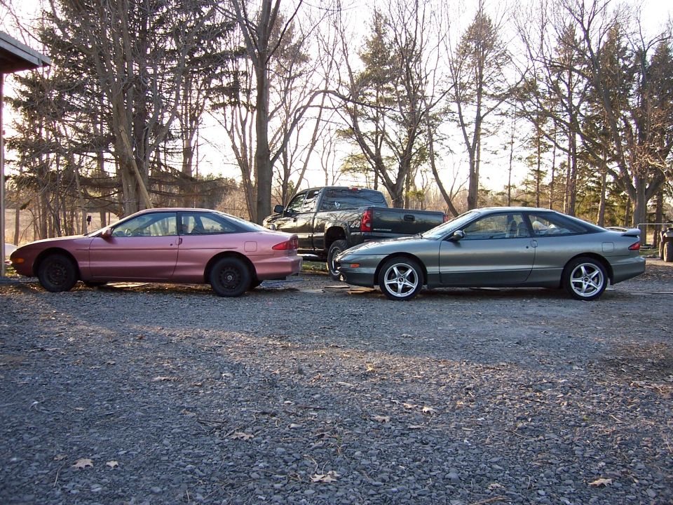 Two Ford Probes, one pink and one gray, parked next to a dark pickup truck