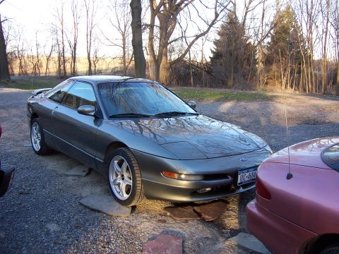 1996 Ford Probe - A gray 1996 Ford Probe is parked on gravel next to another vehicle, with trees in the background.