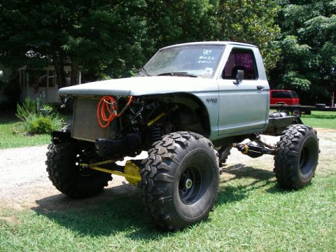 1985 Ford Bronco - This heavily modified pickup truck features oversized tires and a lifted suspension, with '90' written on the windshield.