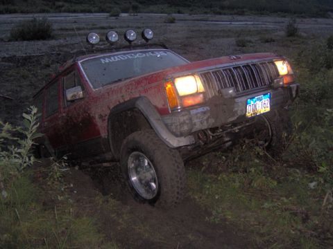 1988 Jeep Cherokee - A red 1988 Jeep Cherokee is stuck in the mud with its headlights on.