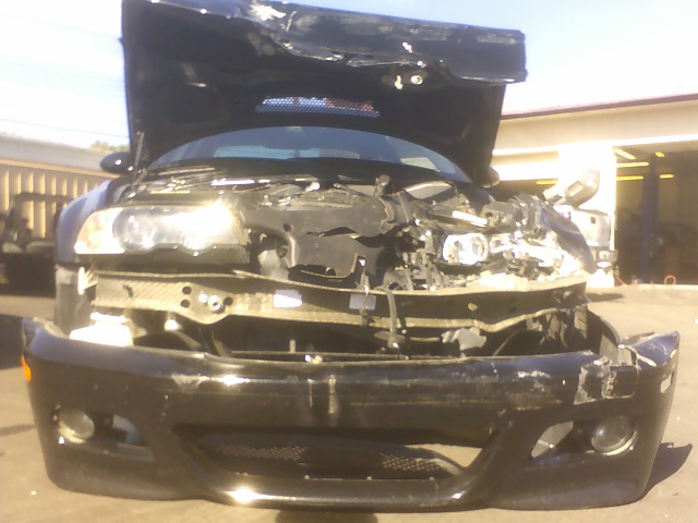 Front-crash-damaged black BMW with hood up, smashed bumper cover hanging, headlights and radiator exposed