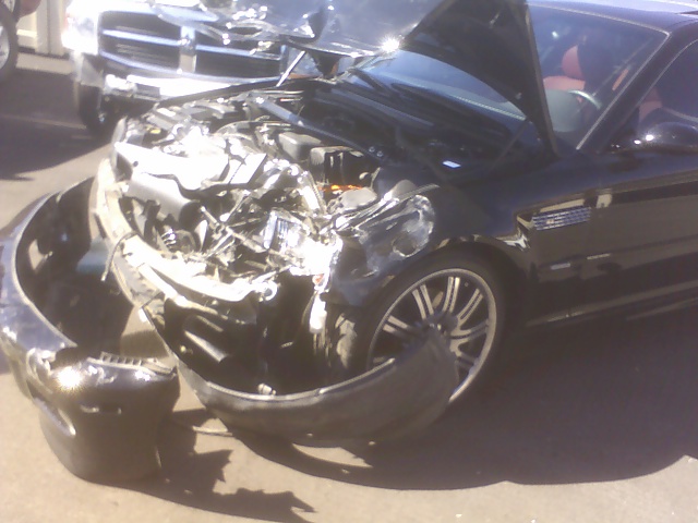 Black BMW with severe front collision damage, bumper cover on ground, chrome multi-spoke wheel, red interior