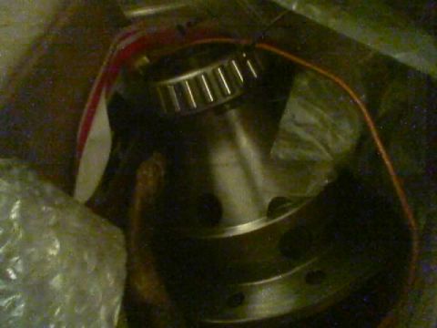 1990 Ford Bronco - tapered roller bearing rests atop a machined steel flanged hub with bolt holes, surrounded by bubble wrap and a striped wire in low light.