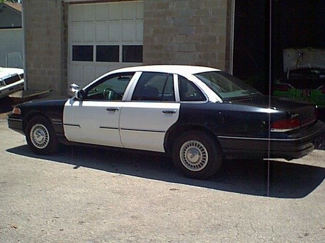 Side view of a 1994 Ford Crown Victoria police car, two-tone black and white, parked in front of a garage