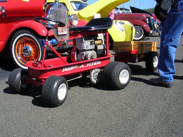 Red Radio Flyer wagon with engine and trailer, parked behind vintage cars