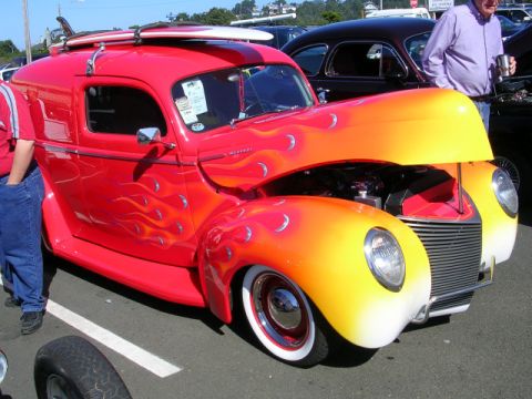 1996 Ford Probe - A custom red panel van with flame decals and surfboards on its roof is displayed at a car show.