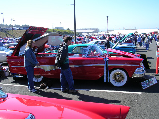Red classic car with hood and trunk open, white wall tires, gold trim. People standing nearby. Other cars and crowd in background