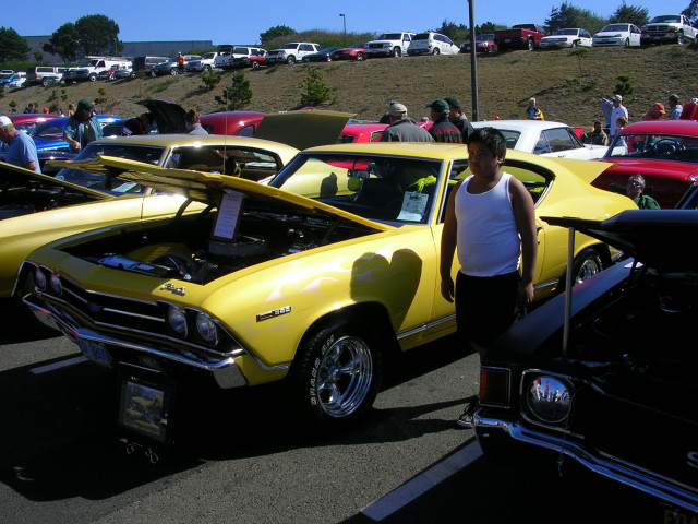 Yellow 1969 Chevrolet Chevelle SS with hood open, parked at a car show. A boy stands next to it
