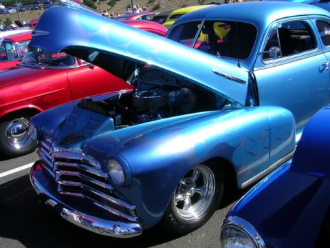 1996 Ford Probe - A custom blue hot rod with flame graphics and its hood open is displayed at a car show, alongside other vehicles.