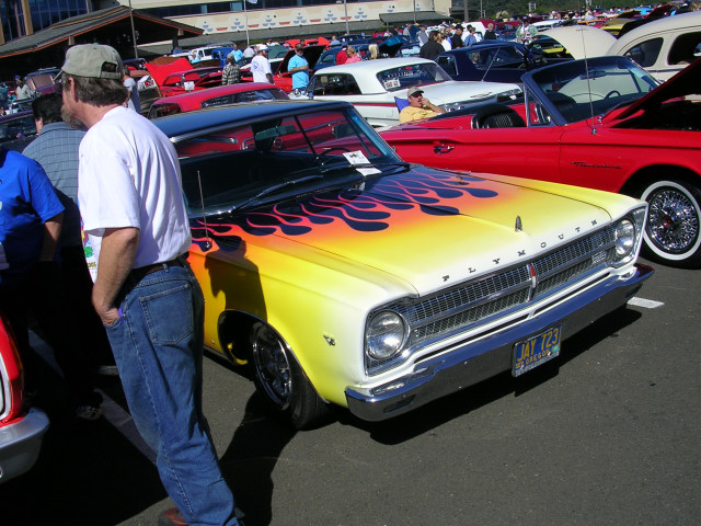 Front view of a 1965 Plymouth Belvedere with yellow and orange flames painted on the hood and fenders. Other cars are visible in the background