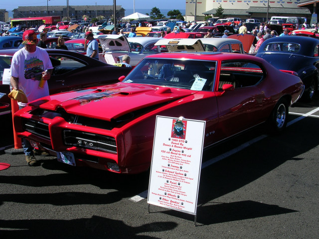 Red 1969 Pontiac GTO with black racing stripes on the hood, parked at a car show