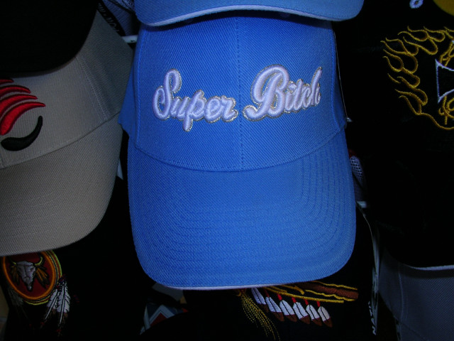 Blue baseball cap with white and silver text that says "Super Bitch". Other hats are visible