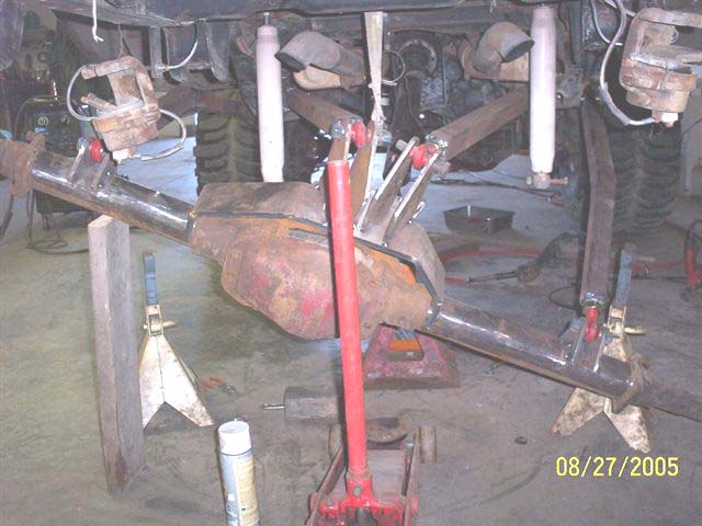 Rear axle assembly on jack stands, with suspension links and shocks attached. Welder and spray can visible