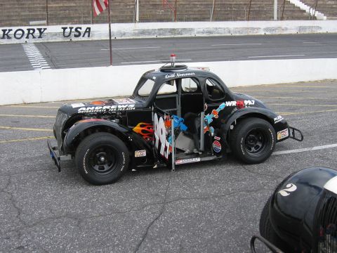 1985 Ford LTD Crown Victoria - A modified stock car with racing decals sits on the asphalt at a speedway, with another vehicle's helmet visible in the foreground.