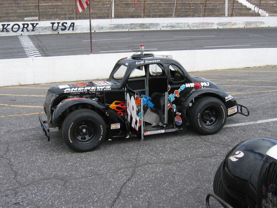 Modified stock car with flames and number 99 on the side, parked at a racetrack. Another car's helmet is in the foreground