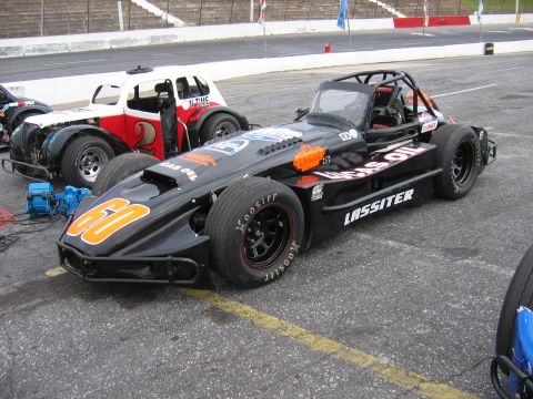 1985 Ford LTD Crown Victoria - A black stock car with racing decals and the number 60 is parked on asphalt next to another race car at a speedway.