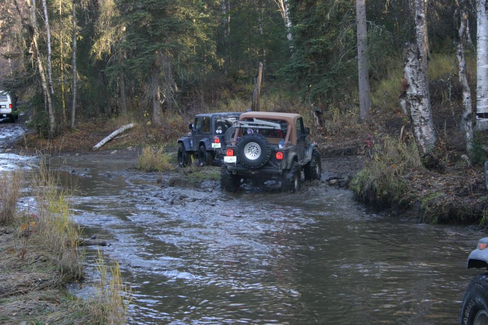 Jeep TJ driving through water, rear view, spare tire visible on back