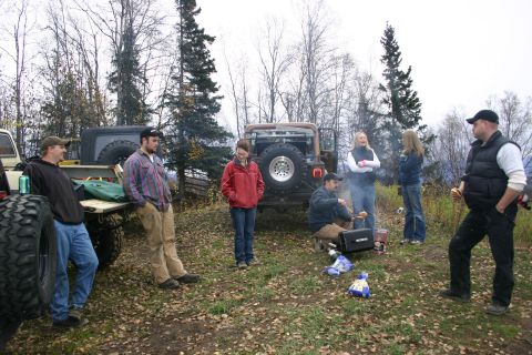 2003 Jeep TJ - A group of people gather around a brown Jeep Wrangler in an outdoor setting, with a second vehicle visible to the left.