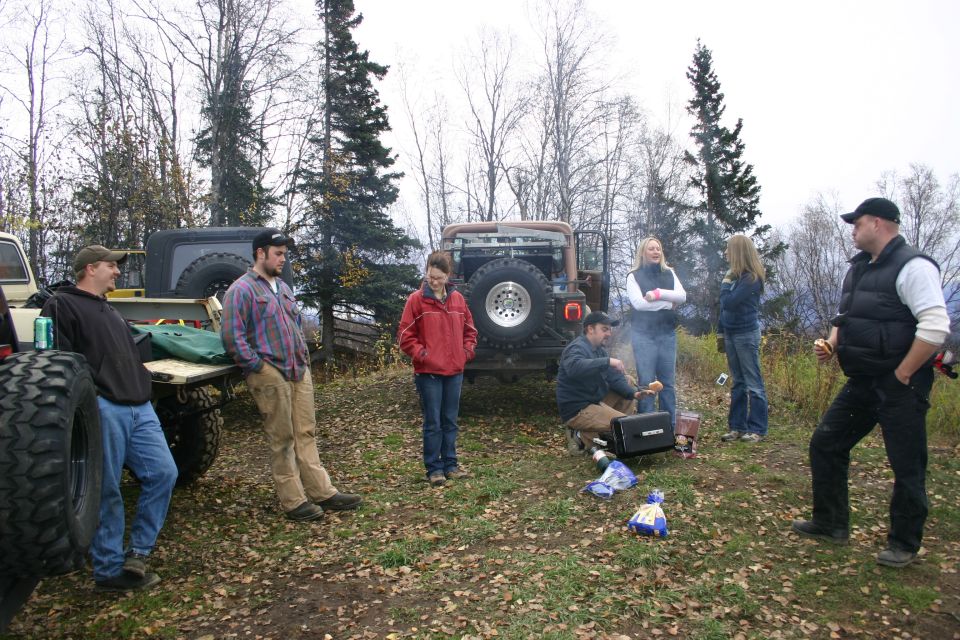 Brown Jeep Wrangler with spare tire on rear, parked outdoors with people