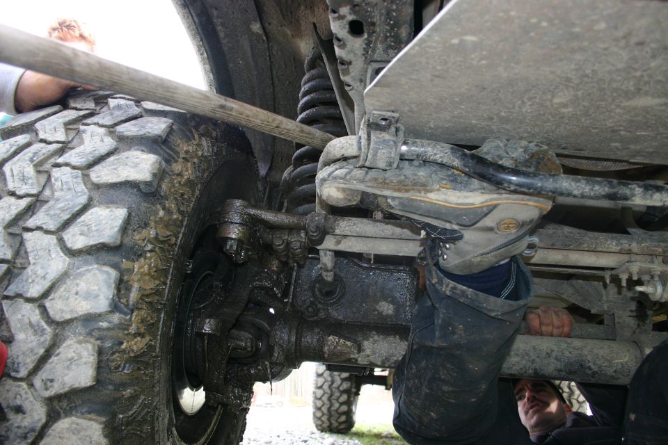 Close-up of a muddy tire and suspension components of an off-road vehicle, with a person's legs visible working underneath