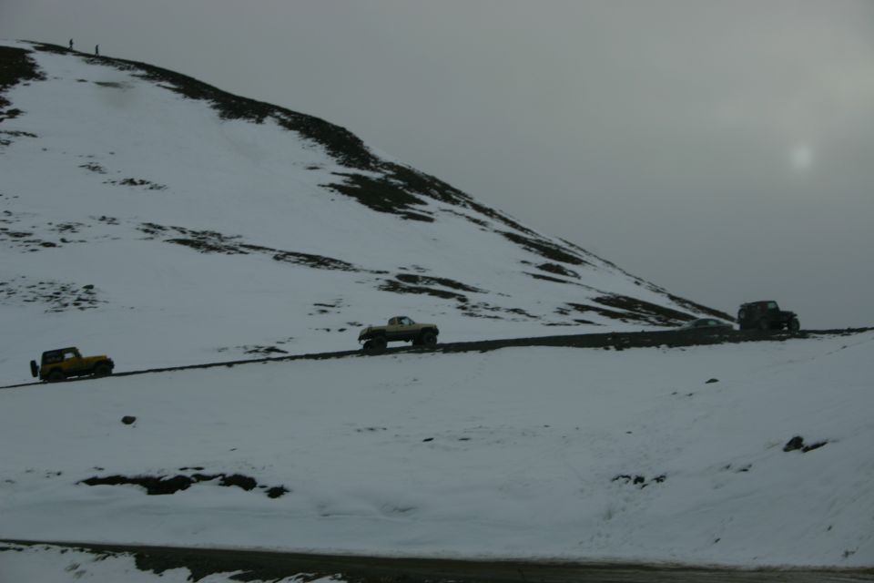 Three off-road vehicles drive along a narrow road on a snow-covered mountain under a cloudy sky