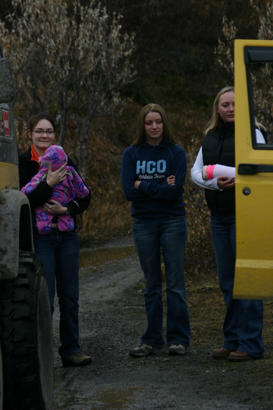 Woman holding a baby, another woman with arms crossed, and a third woman holding a pink object stand near a yellow vehicle door