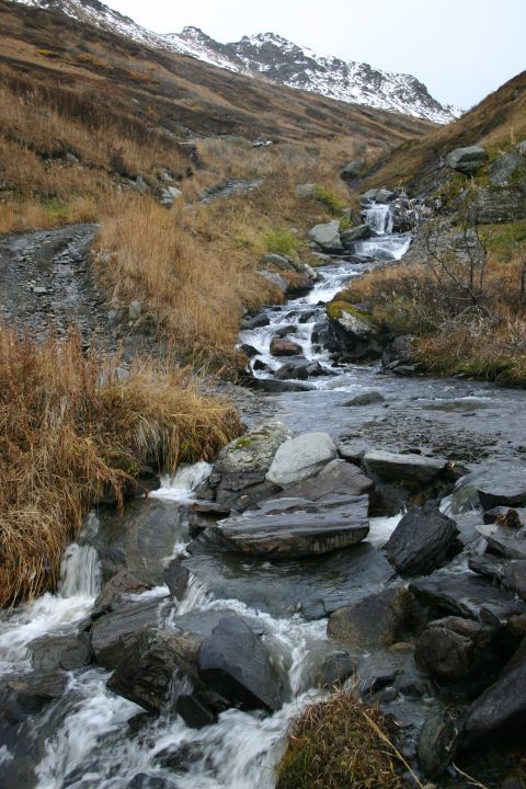 2003 Jeep TJ - A rocky mountain stream cascades down a slope, surrounded by dry vegetation and a backdrop of snow-dusted peaks under an overcast sky.