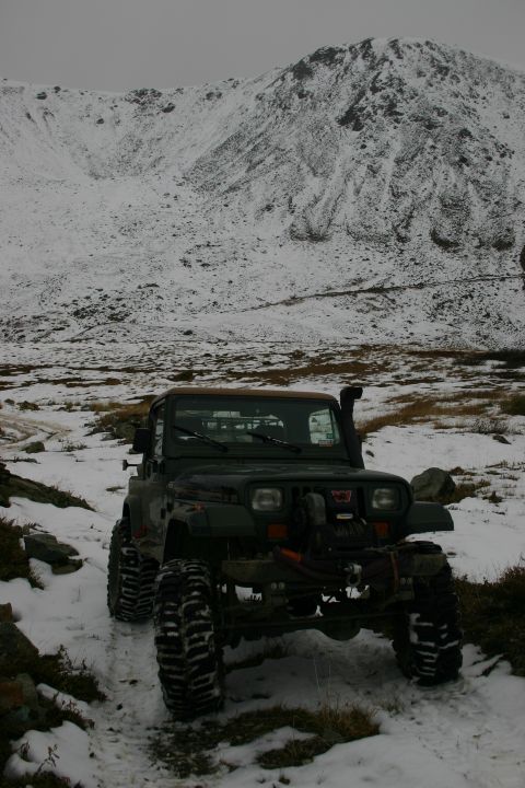 2003 Jeep TJ - A dark green 2003 Jeep TJ navigates a snowy mountain trail, equipped with large off-road tires and a snorkel.