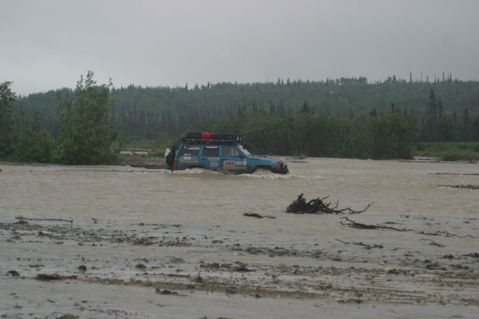 Blue expedition wagon with roof rack fording a wide muddy river by forest