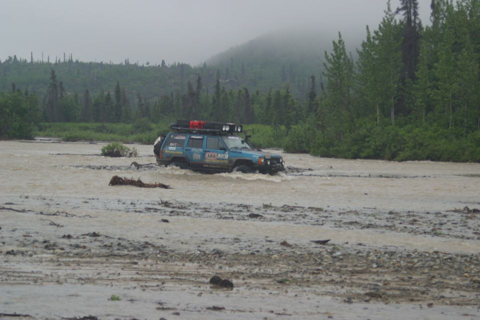 Blue Jeep Cherokee XJ with roof rack and lights driving through shallow muddy water, trees and fog in background