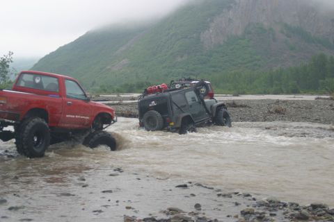 2003 Jeep TJ - Two modified off-road vehicles navigate a shallow, rocky river under a cloudy sky.