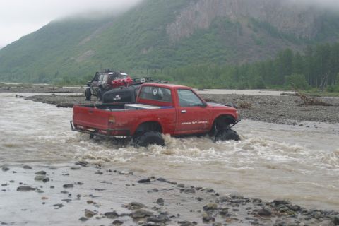 2003 Jeep TJ - Two off-road vehicles navigate a shallow river with rocky banks and a misty mountain backdrop.