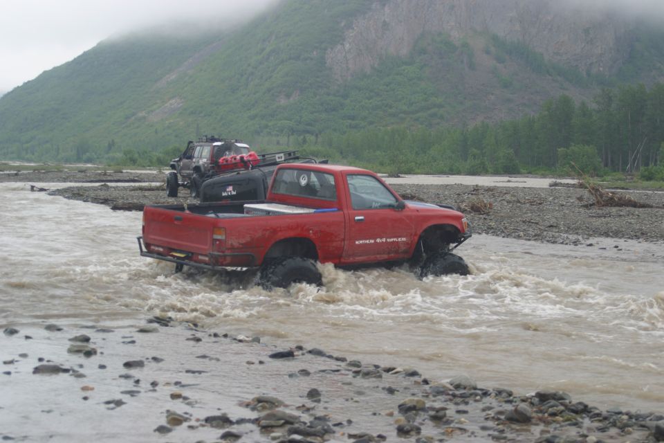 Red pickup truck with large tires driving through a river, with another off-road vehicle behind it