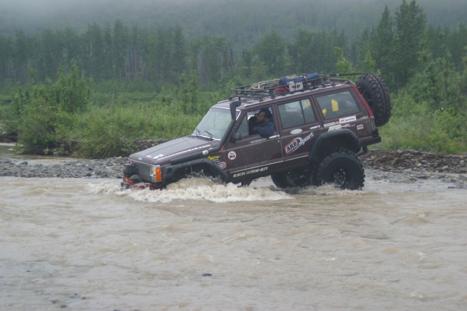 Maroon Jeep Cherokee XJ with roof rack and spare tire crossing a shallow river, driver visible