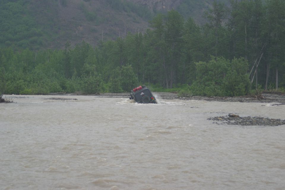 Dark green Jeep TJ driving through a shallow, muddy river with trees in the background