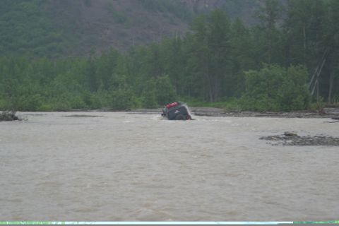 2003 Jeep TJ - A dark SUV navigates through a wide, shallow river, with trees lining the far bank.