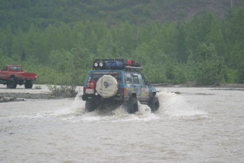 2003 Jeep TJ - Off-road vehicles navigate a shallow river crossing with trees lining the banks.