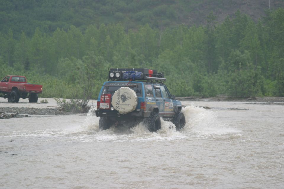 Blue Jeep Cherokee XJ with roof rack and spare tire on rear driving through water, red pickup truck in background