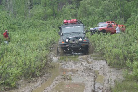 2003 Jeep TJ - A dark green Jeep Wrangler navigates a muddy trail with other vehicles and people in the background.