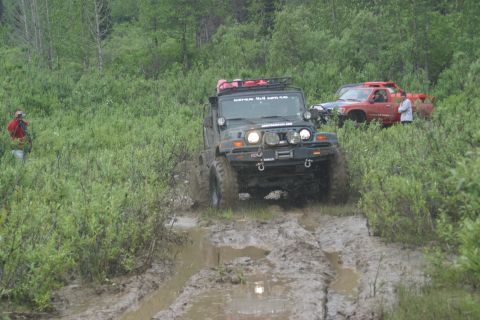 2003 Jeep TJ - A Jeep Wrangler tackles a challenging muddy off-road course, with another vehicle visible in the background.