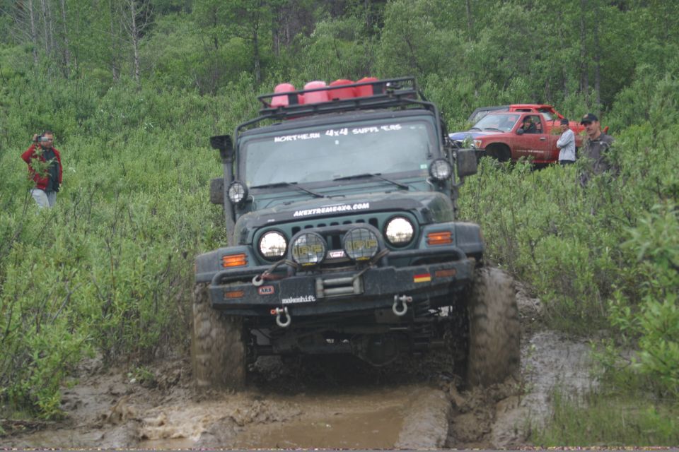 Green 2003 Jeep TJ driving through mud, with a red Toyota Tacoma behind it