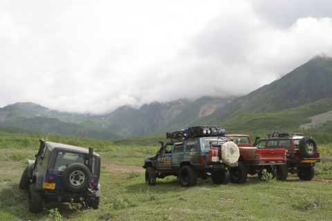 2003 Jeep TJ - A blue 2003 Jeep TJ is parked on a grassy slope with other vehicles nearby, set against a backdrop of cloudy mountains.