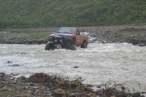 2003 Jeep TJ - Off-road pickup truck navigates a rocky riverbed under overcast skies.