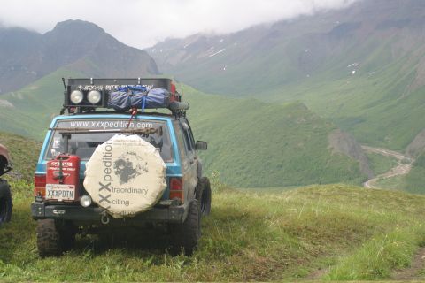 2003 Jeep TJ - An SUV equipped for an expedition is parked on a grassy hillside with mountains and a river in the distance.