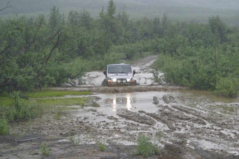 Silver Jeep Wrangler driving through a muddy puddle on a dirt road with trees on either side