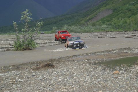 2003 Jeep TJ - Two off-road vehicles navigate a rocky riverbed, with a red truck following a silver SUV through the water.